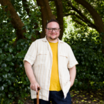 Elliot, a fat, non-binary white person is holding a wooden cane and wearing a yellow t-shirt and a linen button-down shirt, and olive colored-rectangular glasses. They are smiling and standing in front of big, green leafy trees in Golden Gate Park in San Francisco.