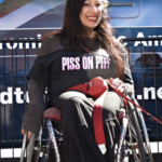 Una mujer latina con pelo negro largo lleva una camiseta que dice “Piss on Pity” (Mearse en la Lastima) y mira directamente a la cámara con una gran sonrisa y labial rojo. Está sentada en su silla de ruedas frente a una cerca durante una protesta o acción directa. Hay un vehículo grande detrás de ella con letras ilegibles en un costado.