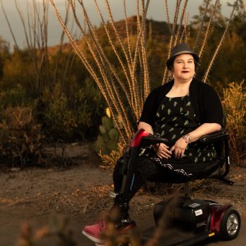 Naomi Ortiz, a light-skinned mestizx, with dark hair and silver hoop earrings, sits in their scooter in front of an ocotillo in the Sonoran desert. They are wearing a fedora hat, hoop earrings, black dress with cacti print, textured tights, silver bracelet, and pink boots. In the distance is a mountain and cacti.