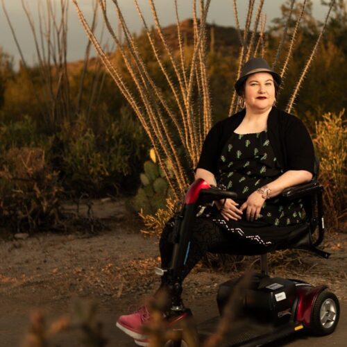 Naomi Ortiz, a light-skinned mestizx, with dark hair and silver hoop earrings, sits in their scooter in front of an ocotillo in the Sonoran desert. They are wearing a fedora hat, hoop earrings, black dress with cacti print, textured tights, silver bracelet, and pink boots. In the distance is a mountain and cacti.