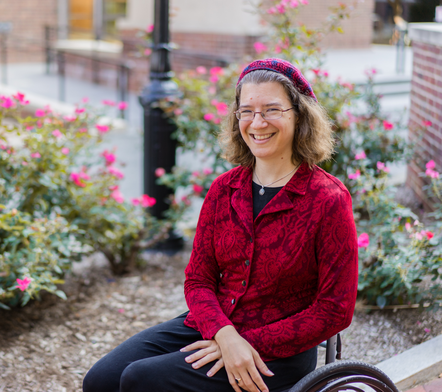 Julia Watts Belser, a white Jewish woman with curly brown hair, sits happily in her wheelchair in front of a pink flowering bush. She's wearing a patterned red blazer and red kippah (beret) to match.