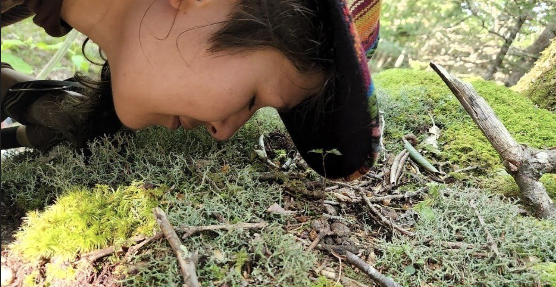 A photo of Marina, nose to moss. The feeling is intimate: a diverse community of mosses, with various textures and leaf patterns, cover the forest floor. It is daylight. Just above the surface of the earth is a human face in profile, head parallel to the ground, eyes closed, inhaling and gently smiling. She is wearing a colorful cap, whose rim is dark and in shadow. Against the rim of the cap -towards the camera lens - is a small 2-leaf sprout growing upwards, a future tree. In the distance are blurry trees pierced by light, suggesting the presence of a larger forest.