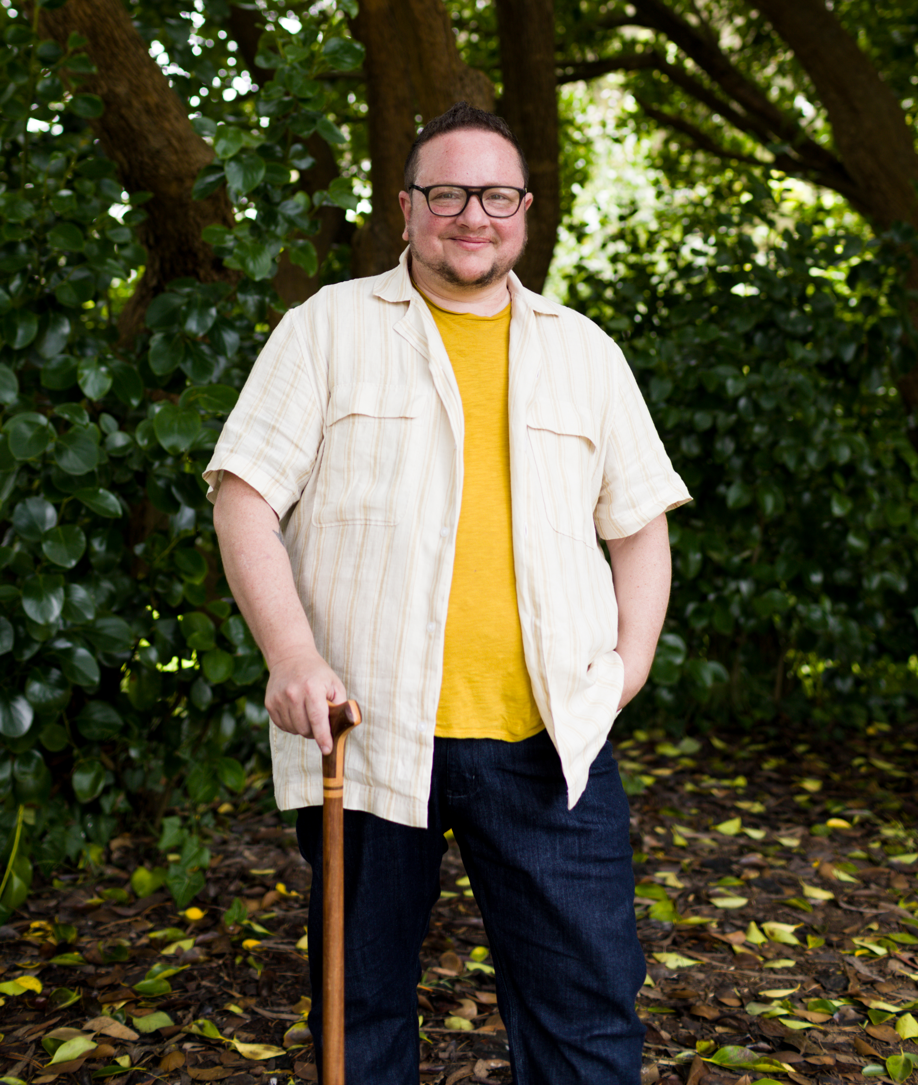 Elliot, a fat, non-binary white person is holding a wooden cane and wearing a yellow t-shirt and a linen button-down shirt, and olive colored-rectangular glasses. They are smiling and standing in front of big, green leafy trees in Golden Gate Park in San Francisco.