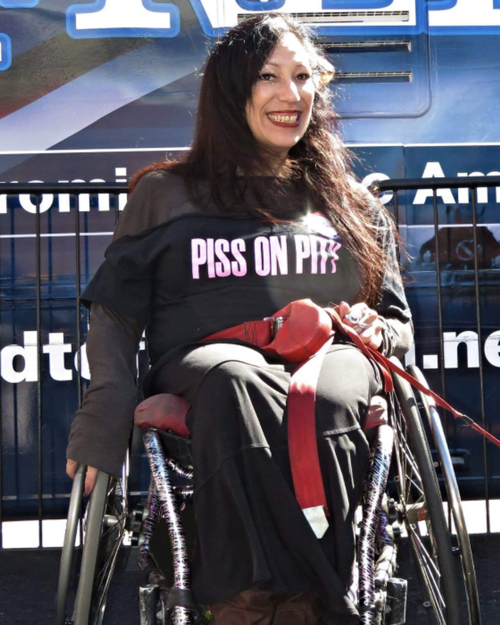 Una mujer latina con pelo negro largo lleva una camiseta que dice “Piss on Pity” (Mearse en la Lastima) y mira directamente a la cámara con una gran sonrisa y labial rojo. Está sentada en su silla de ruedas frente a una cerca durante una protesta o acción directa. Hay un vehículo grande detrás de ella con letras ilegibles en un costado.